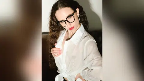 Stylish woman with curly hair wearing black glasses and a white button-down shirt in a moody indoor setting.