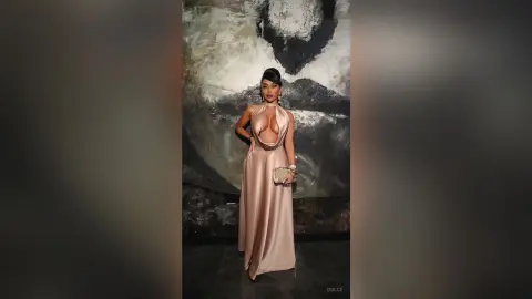 Dolce poses in a long, tan cut-out dress that shows cleavage and her midriff against a dark, textured wall.