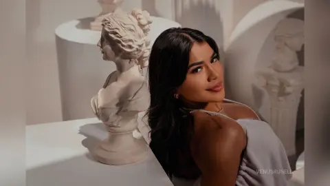 Young woman in a grey top posing elegantly next to a white classical marble bust in a bright studio setting.