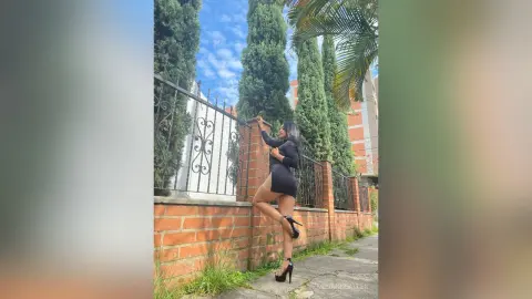Stylish woman in a black mini dress and high heels posing by a decorative wrought iron fence outdoors.