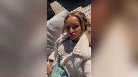 Blonde woman in a white fuzzy coat relaxing outdoors on a sunny day.