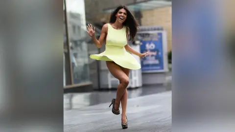 Cheerful woman in a pale yellow mini dress dancing on a city street in high heels.