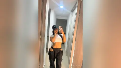 Young woman in white crop top and black cargo pants taking a mirror selfie in a modern hallway.