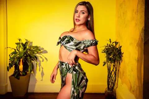 NinaBonetti poses in a green and white tropical print off-the-shoulder top and matching skirt with a high slit against a bright yellow wall.