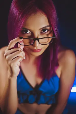 Young woman with pink hair wearing glasses and a blue crop top in moody, cinematic lighting.
