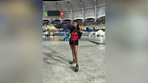 AmyJacobs stands in a large stadium wearing a red corset top and black shorts with black boots.