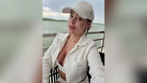 Smiling woman in a white textured outfit and baseball cap relaxing by a scenic waterfront view.