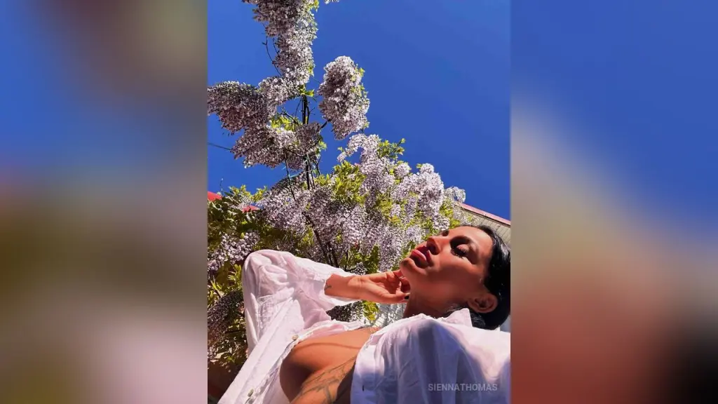 SiennaThomas β Stylish woman in a white linen shirt relaxing under blooming lilac trees against a bright blue sky.