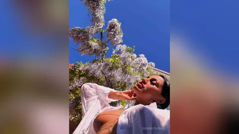 Stylish woman in a white linen shirt relaxing under blooming lilac trees against a bright blue sky.