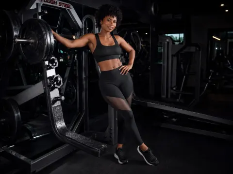 KylieDecks — KylieDecks poses in a black sports bra and leggings while leaning against weightlifting equipment in a gym.