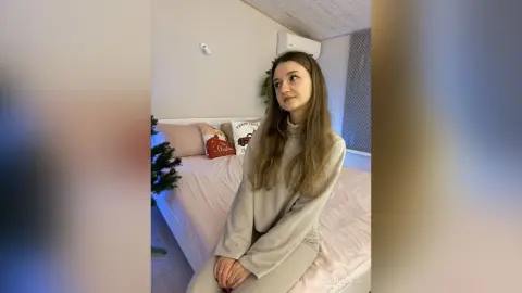 Young woman in a beige turtleneck sweater sitting on a bed in a bright, cozy bedroom.