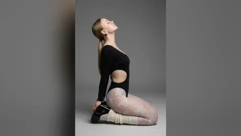 SophiePeachy poses on her knees against a gray background, wearing a black cutout bodysuit and white patterned tights.