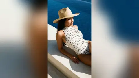 Woman in floral white swimsuit and straw sun hat relaxing by a sunny blue swimming pool.