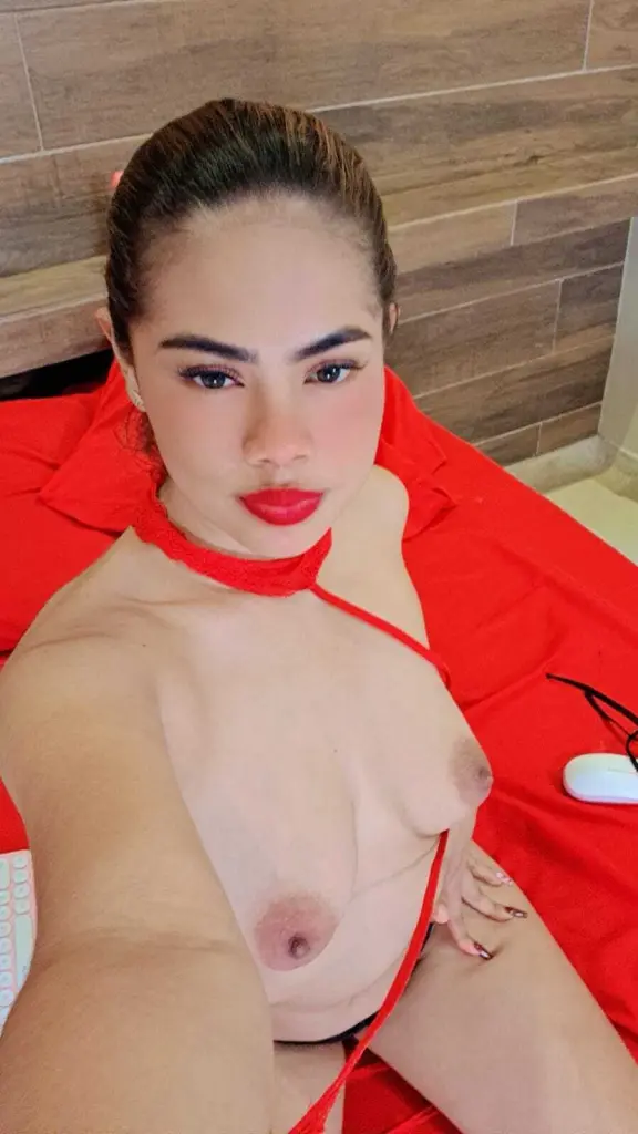 LorenaVargass poses in a red lace top, looking off-camera with her dark hair pulled back in a bun against a grey wall.