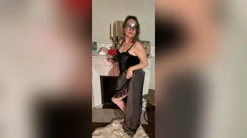 Woman in black lace lingerie and silver masquerade mask posing in a luxury living room.