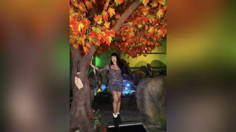 Young woman in a leopard print mini dress posing by an autumn tree in a moody, dimly lit indoor setting.
