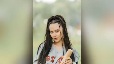 Young woman with long dark hair in a casual grey sweatshirt posing outdoors in soft natural light.
