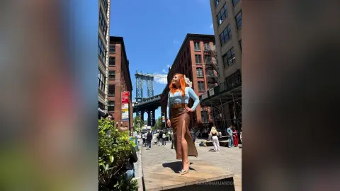 Redheaded woman in a blue top and brown skirt standing on a sunny city street near a bridge.