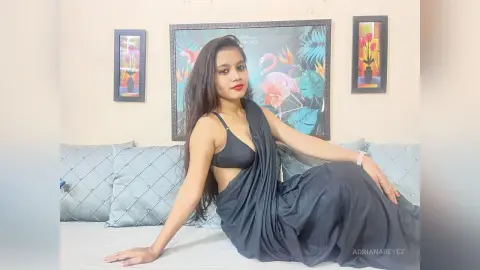 AdrianaReyez poses sitting on a light blue couch, wearing a black sleeveless top and a long black skirt with her long dark hair down.