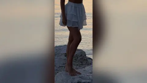 Woman in a white tiered mini skirt standing on coastal rocks by the ocean at sunset.
