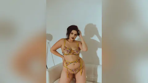 Brunette woman in floral print bikini posing confidently against a white wall in a minimalist indoor setting.