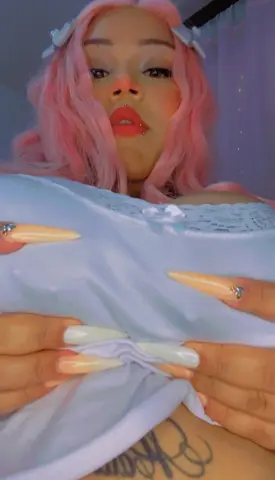 Woman with pink hair and long manicured nails wearing a light blue lace top in a soft, dreamy setting.