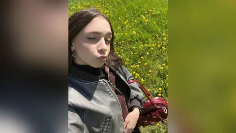 Young woman in a grey denim jacket and black turtleneck posing in a sunny green meadow with wildflowers.