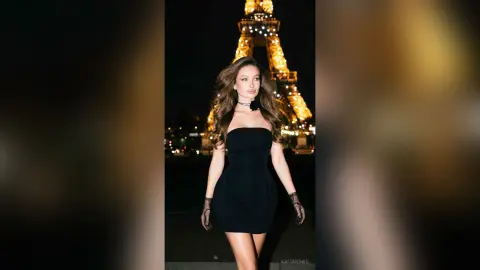 KatyMonro poses in a short black strapless dress and black gloves with the lit Eiffel Tower behind her at night.