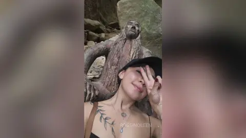 RossiNoleen smiles while posing outdoors against a stone statue, showing a leaf tattoo on her shoulder and chest.