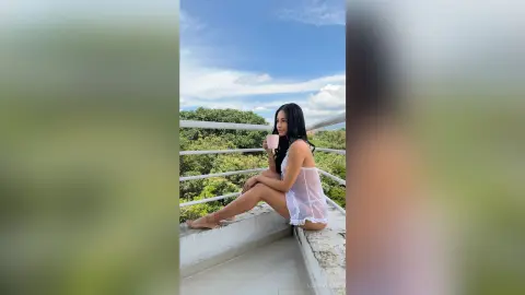 Relaxed woman in a white summer dress sipping coffee on a sunny balcony overlooking lush green trees.