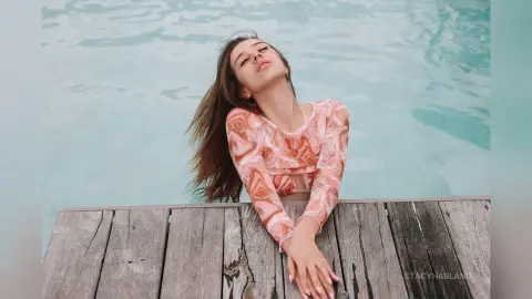 StacyHarland leans back over a wooden dock with her eyes closed, wearing a long-sleeved peach patterned top against a background of blue water.
