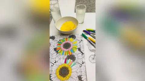 Colorful hand-drawn sunflower illustration on a sketchbook next to markers and a bowl of yellow food on a white table.