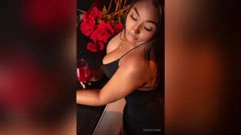 NaiomiLanne — Glamorous woman in a black dress holding a red drink near red roses in moody lighting.