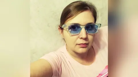 Woman with brown hair wearing blue-tinted sunglasses and a pink t-shirt in a bright indoor setting.