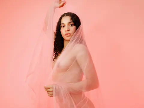Young woman with curly hair draped in sheer pink fabric against a soft pink background for a dreamy portrait.