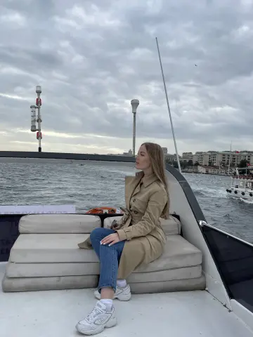 Blonde woman in a beige trench coat and jeans relaxing on a boat deck under a cloudy sky.