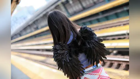 Woman with black feathered wings standing on a city subway platform in a dreamy urban setting.