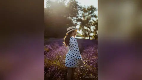 Stylish woman in a polka dot dress and straw hat standing in a dreamy purple lavender field at sunset.