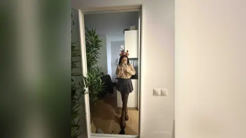 Young woman in a beige sweater and black skirt taking a mirror selfie in a modern minimalist room.