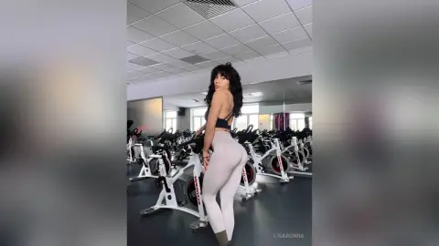Athletic woman in black sports bra and grey leggings posing in a bright modern indoor cycling studio.