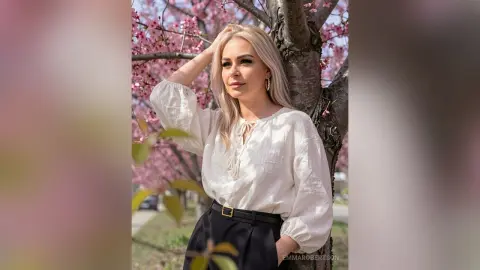 Blonde woman in a white linen blouse posing by blooming pink cherry blossom trees in a serene spring setting.