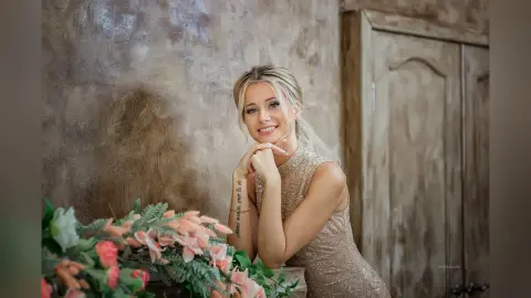 Blonde woman in a sparkling gold sequin dress posing thoughtfully near pink flowers in a rustic room.