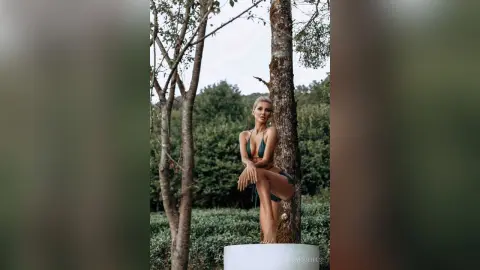 Blonde woman in a green bikini posing gracefully by a tree in a lush, serene forest setting.