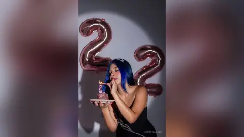 Young woman with blue hair in a black dress blowing out a candle on a birthday cake near rose gold number 22 balloons.