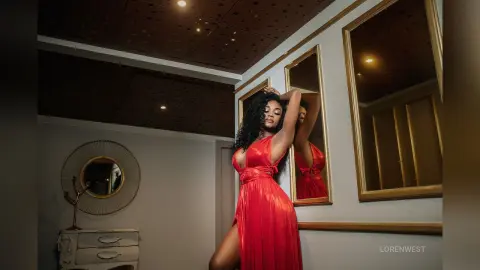 Elegant woman in a glamorous red evening gown posing in a luxury mirrored dressing room with moody lighting.
