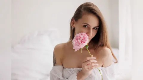 IngridBeckton holds a pink flower to her face, showing her long brown hair and bare shoulders in a white bed setting.