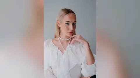 MarryDavies poses in a white ruffled blouse and a pearl necklace, looking directly at the camera with her hand touching her neck.