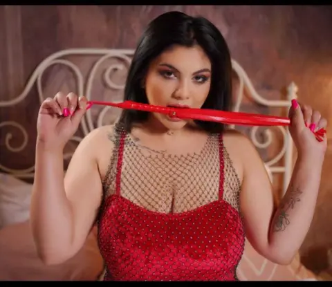 Woman in a red sequin dress holding a red prop in a bedroom setting with a playful mood.