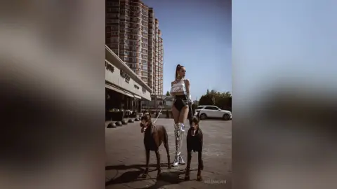 Stylish woman in a white crop top and silver boots walking two large dogs in an urban city setting.