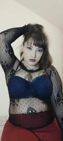 Woman in black lace mesh top and blue bra posing with a moody expression against a white wall.